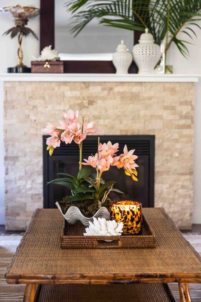 A view of a woven rattan serving tray on a wooden coffee table in front of a fireplace, holding a silver shell-shaped bowl with artificial pink orchids, a glass leopard print candle, and a white coral cluster.