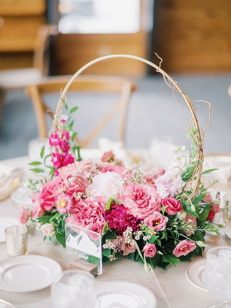A monochromatic pink wedding centerpiece featuring a gold hoop with vine accents framing a dense cluster of pink peonies and roses on a neutral tablescape.