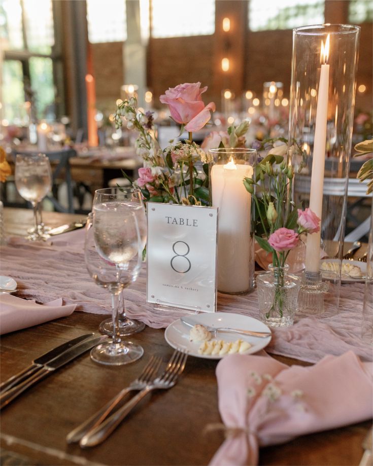 An industrial glam wedding tablescape featuring pink roses in bud vases, a mauve gauze runner, and tall taper candles on a rustic wood table.