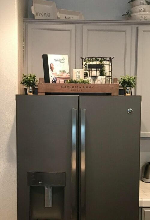 A wooden Magnolia Home crate provides top of the fridge organization for a cookbook and wire basket. This is a functional and aesthetic top of the fridge storage solution.