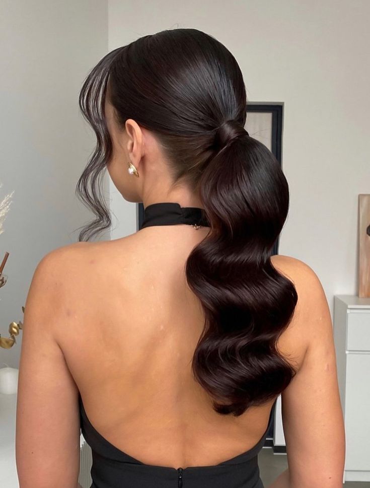 Back view of a woman with dark hair styled in a low ponytail featuring deep, uniform "S" waves. She is wearing a black halter-neck dress.