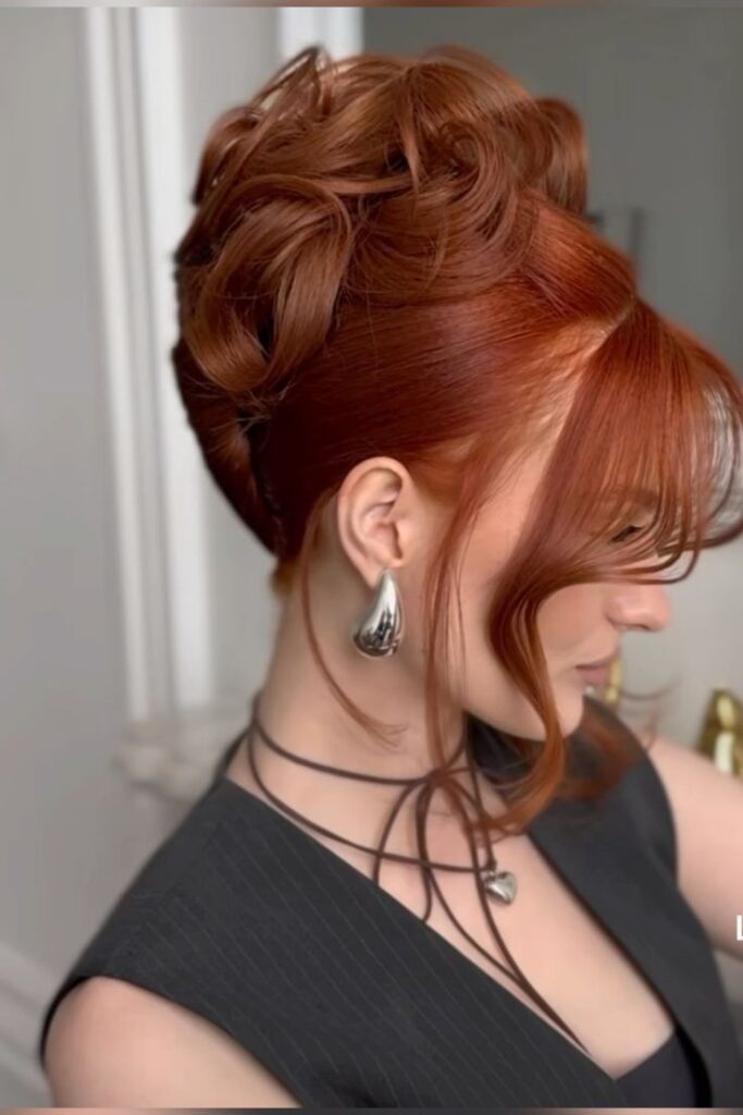 A side view of a woman with vibrant red hair styled in a high, sculptural updo with large, glossy barrel curls pinned to the top of the head.