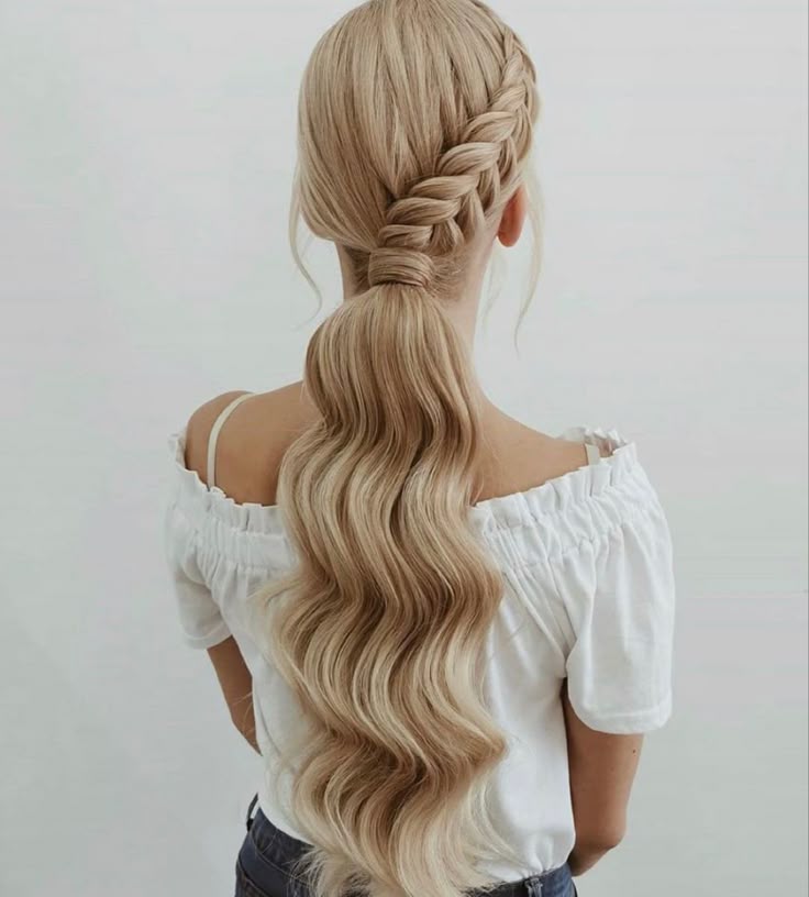 A back view of a person with long blonde hair featuring a large diagonal braid that merges into a low ponytail with wide, flowing waves.