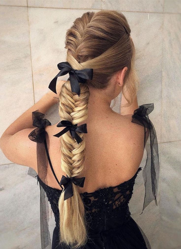 A back view of a person with blonde hair in a long fishtail braid adorned with three small black ribbons along the length.