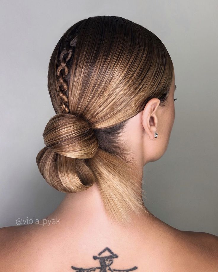 A back view of a person with light brown, glossy hair featuring a central vertical braid leading into a structured low bun with a sleek, fanned-out tail.