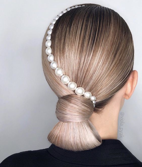 A back view of a person with ash-blonde hair in a smooth low bun, decorated with a curved row of white pearls of increasing size along the side of the head.