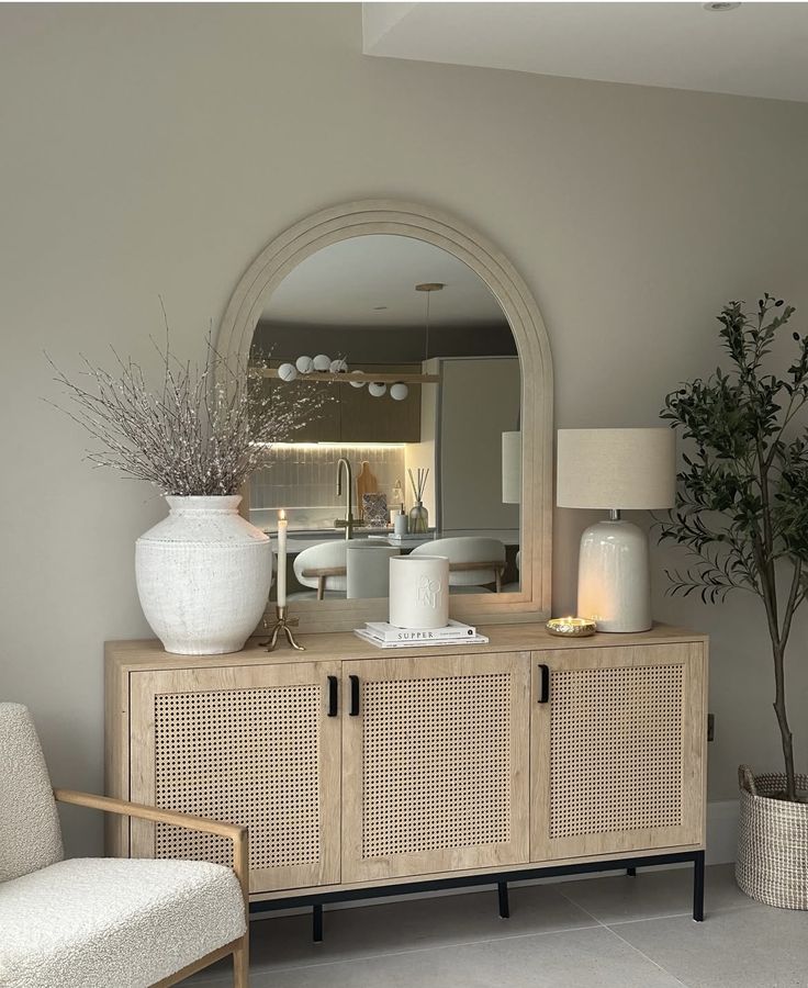 A light oak cabinet with black handles situated under a large arched mirror, featuring a textured white vase with thin branches and a white table lamp.