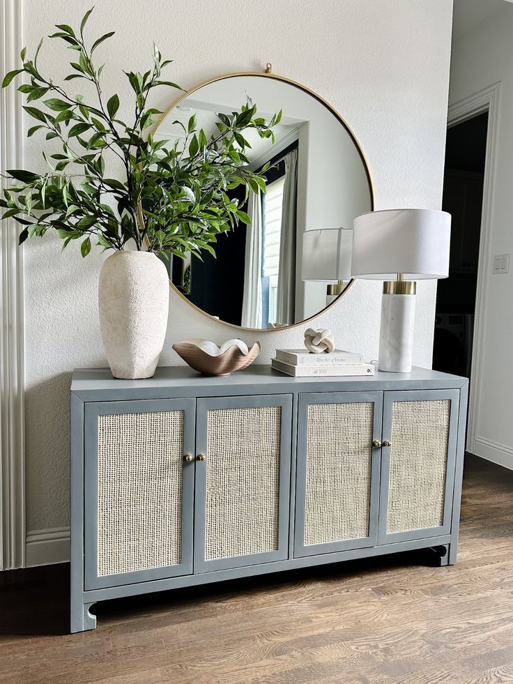 A sage green four-door console table with woven panels, decorated with a marble table lamp, a round mirror, and a large white vase filled with leafy green branches.