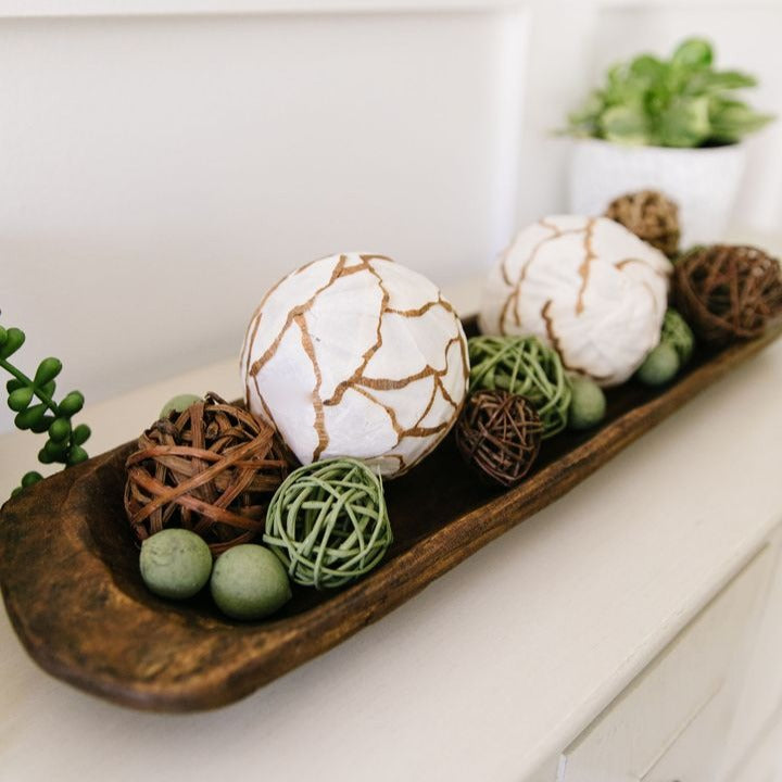 A dark, weathered long wooden dough bowl resting on a white dresser holds decorative orbs, including two white textured ones with brown pattern, three dark wood twig spheres, three small green string-wrapped balls, and three small smooth green stones.