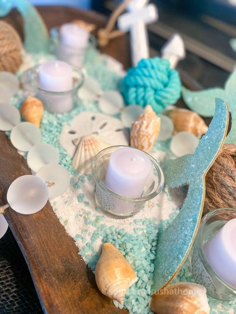 A dark wood tray filled with white sand and turquoise pebbles, accented by seashells, sand dollars, and sea glass. Small glass votive candles and a glittery teal mermaid tail cutout complete this summer dough bowl look.