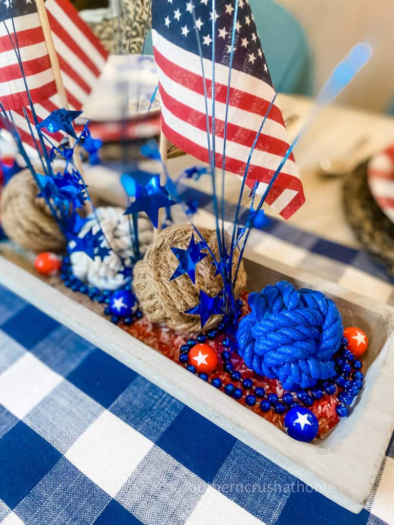 A side angle view of a rectangular wooden dough bowl filled with small red rocks, blue beaded garland, large rope balls, blue and red star sprays, and small American flags, placed on a blue and white buffalo plaid tablecloth.