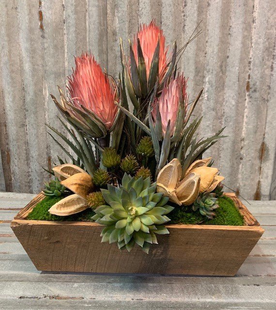 A rectangular wooden summer dough bowl decor piece containing vibrant green succulents, dried pods, and tall, pink-tipped protea flowers set in green moss. It serves as a textured, rustic decor focal point for any indoor or outdoor table.