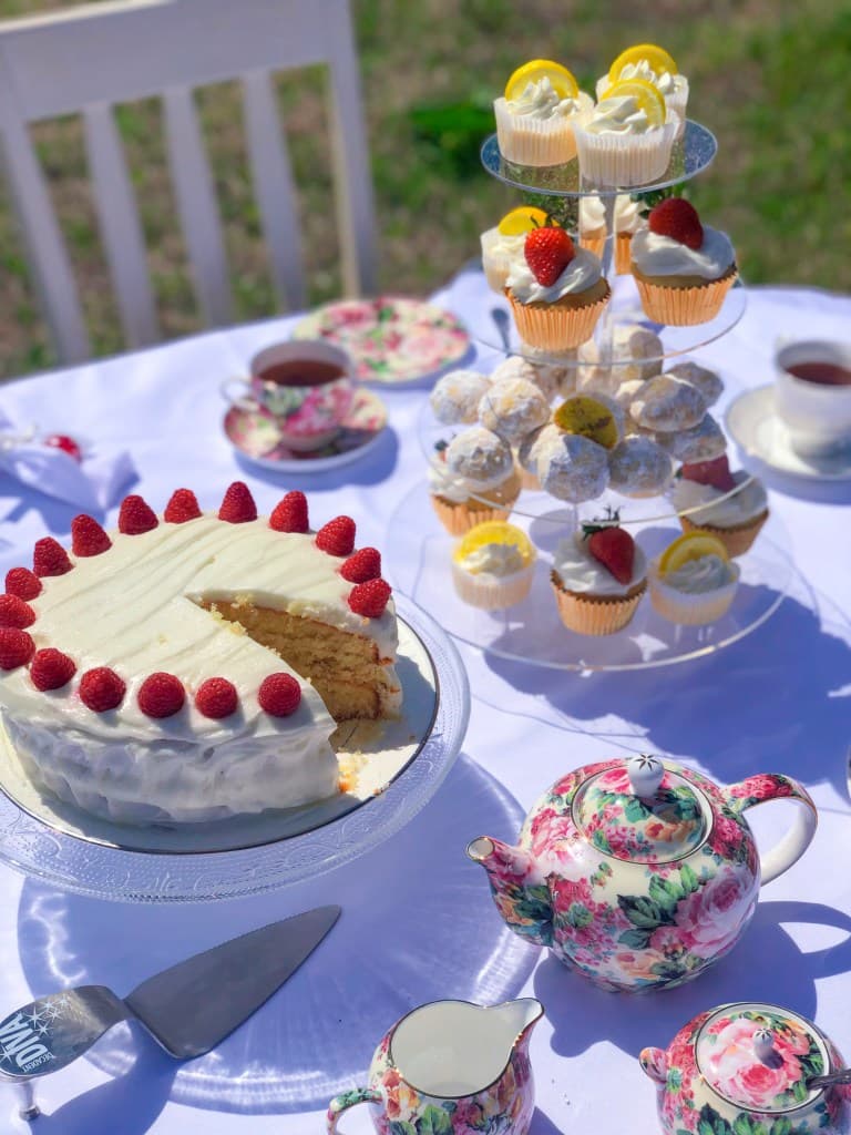 An outdoor tea setting on a white tablecloth featuring a white frosted cake decorated with fresh raspberries, a clear tiered stand with lemon cupcakes, and a pink floral teapot in the sunlight.