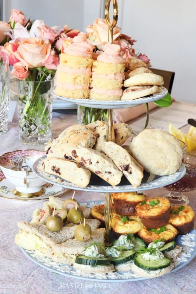 A three-tiered light blue floral tea tray filled with finger sandwiches, quiches, scones, and pink frosted cakes, set next to a vase of peach roses.