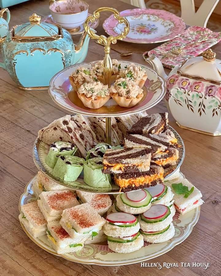 A vintage-style tiered tray stacked with various savory tea sandwiches, wraps, cucumber rounds, and small pastry cups on a wooden table.