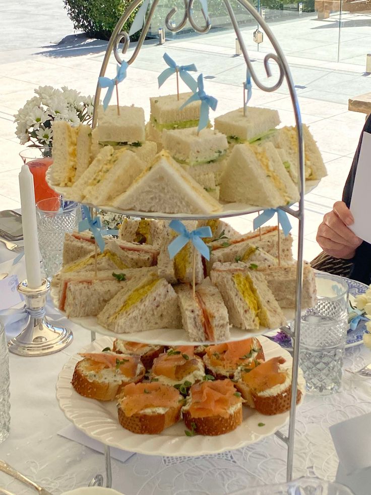 A white three-tiered wire stand holding rows of triangular tea sandwiches and smoked salmon crostini, decorated with small light blue bows.