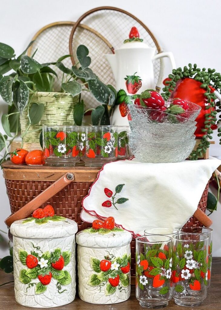 A wicker picnic basket topped with strawberry-patterned glassware, ceramic canisters with 3D strawberry lids, and an embroidered white napkin.