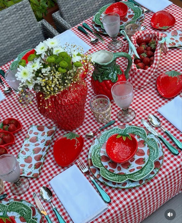 An outdoor table set with a red and white gingham tablecloth, strawberry-shaped ceramic dishes, a large strawberry vase with flowers, and baskets of fresh strawberries.