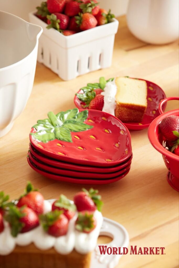 A stack of red ceramic plates shaped like strawberries sitting on a wooden table next to fresh strawberries and a dessert topped with white cream.