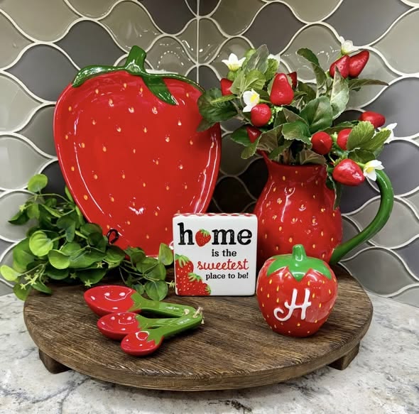 A wooden circular riser holding a large strawberry plate, a red pitcher filled with faux berries, strawberry-themed measuring spoons, and a small decorative sign.b