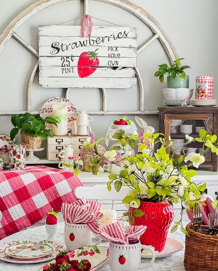 A farmhouse-style dining area featuring a large rustic wooden strawberry sign on the wall, a table set with red gingham linens, strawberry-patterned mugs, and a large strawberry-shaped vase filled with white flowers.
