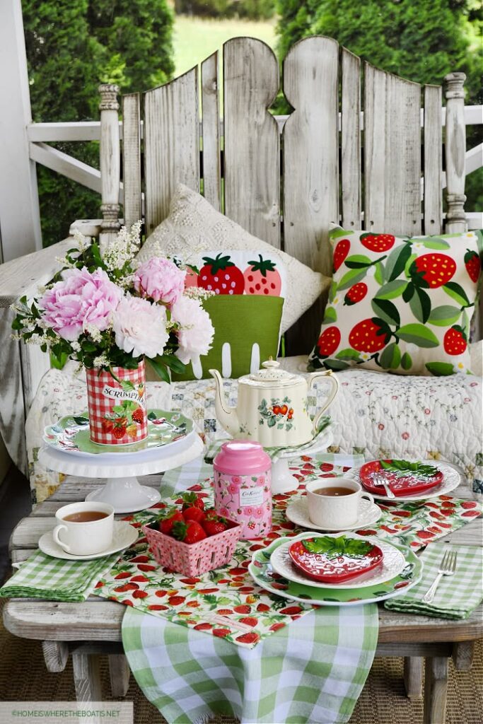 A rustic outdoor wooden table set for tea with strawberry-patterned napkins, plates, and pillows, featuring a teapot and fresh pink peonies.