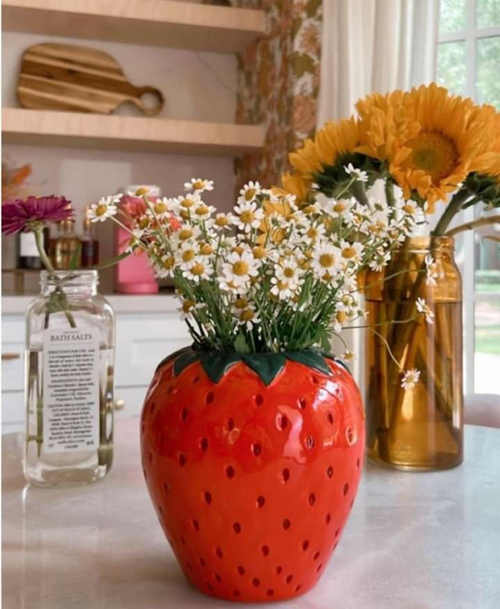 A bright red ceramic vase shaped like a large strawberry filled with small white daisies, sitting on a white marble surface.