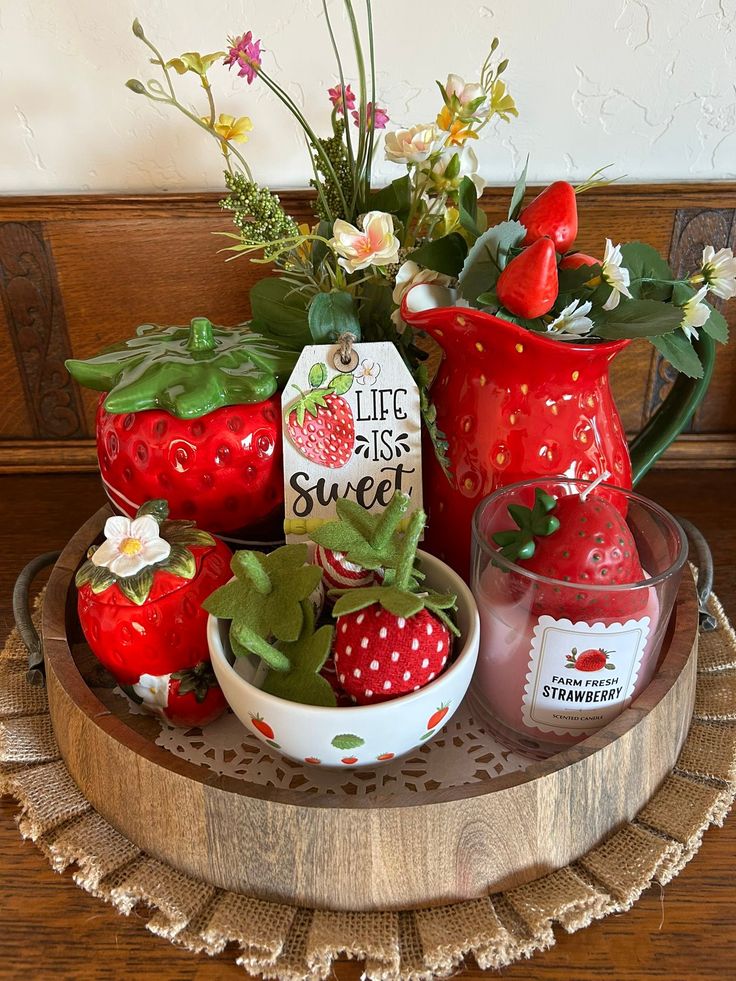 A circular wooden tray holding a strawberry-shaped ceramic jar, a red pitcher filled with flowers, a strawberry-scented candle, and a small bowl of felt strawberries.