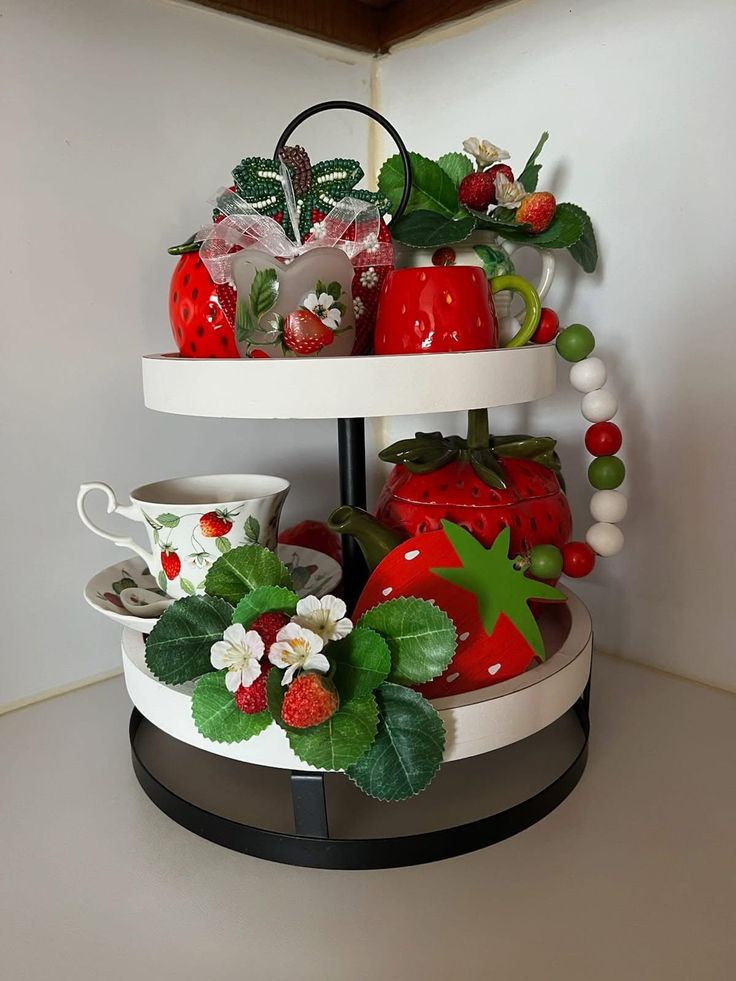 A white and black two-tiered tray displaying a floral strawberry teacup, a large ceramic strawberry pot, a themed teapot, and a red, white, and green beaded garland.