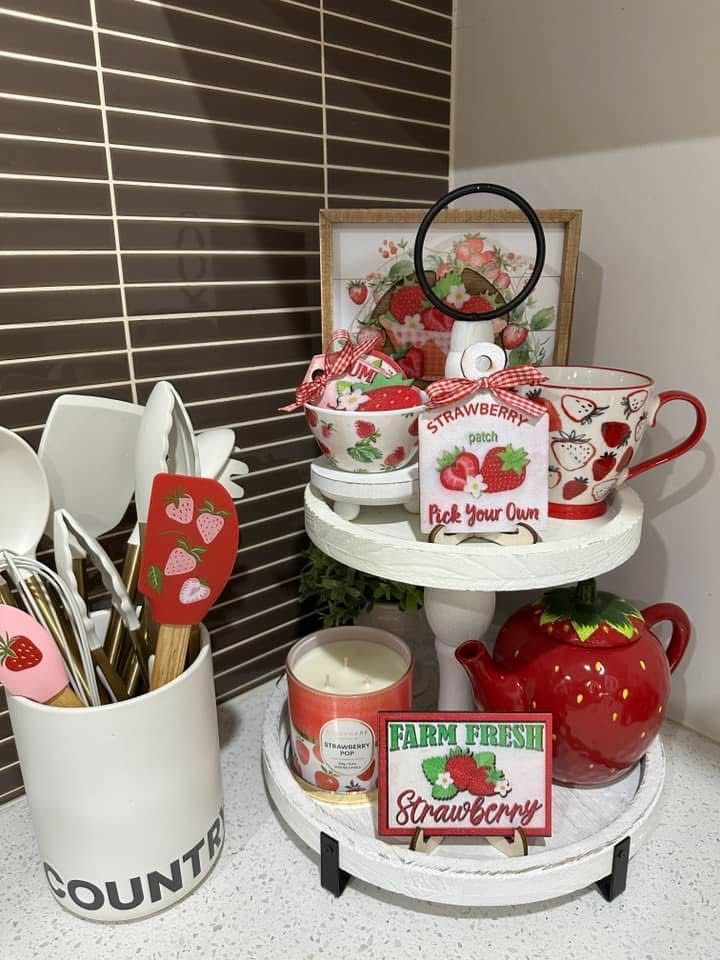 A white two-tiered tray on a kitchen counter featuring a strawberry-shaped teapot, themed mugs, and "Farm Fresh" signs, next to a utensil holder filled with strawberry-patterned spatulas.