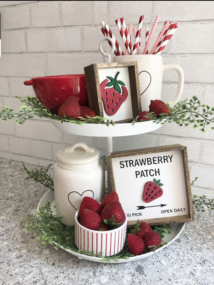 A white two-tiered tray on a granite countertop filled with fresh strawberries, a "Strawberry Patch" wooden sign, a white heart canister, and red and white patterned straws.