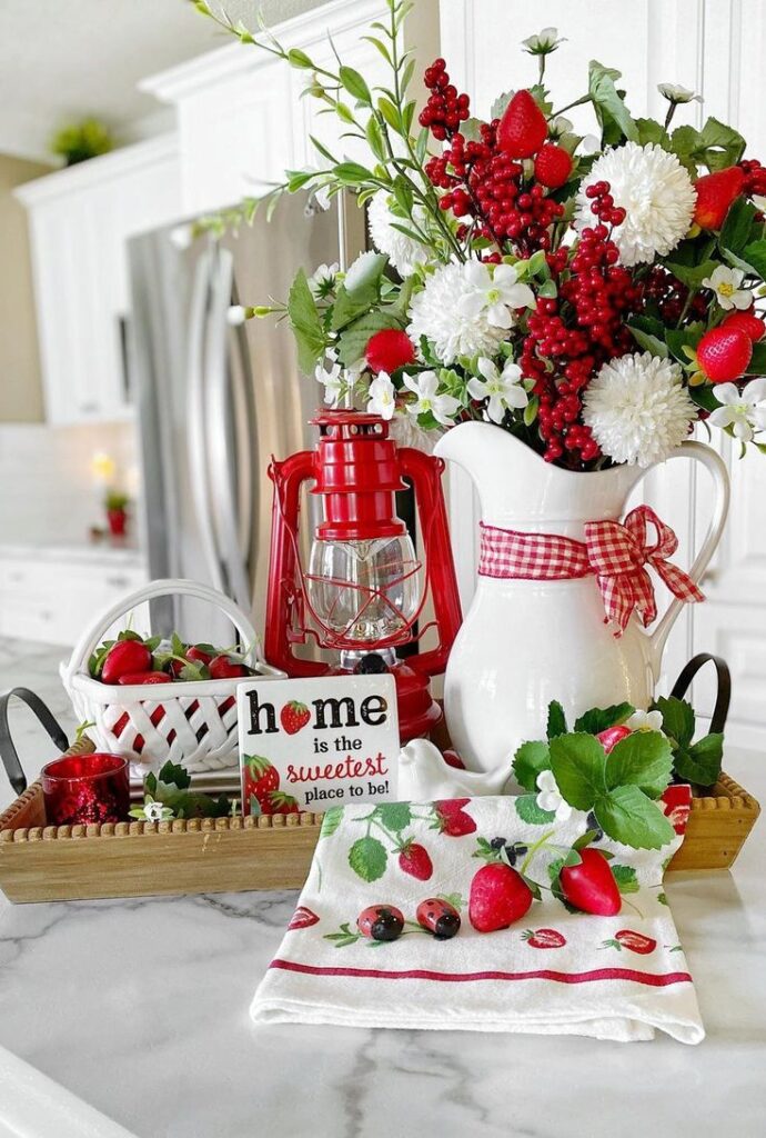A decorative tray on a marble counter featuring a white pitcher with a red gingham bow, a red lantern, a small white basket of strawberries, and a strawberry-themed tea towel.