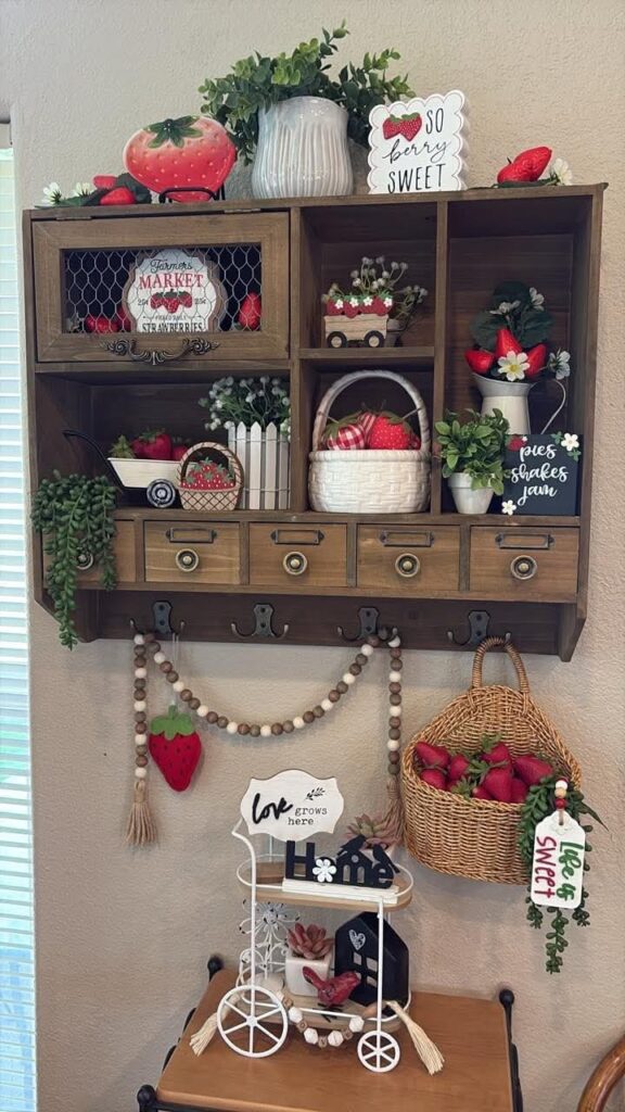 A wooden wall hutch with multiple shelves and drawers filled with strawberry-themed decor, including baskets of berries, a "Farmer's Market" sign, and a beaded strawberry garland hanging below.