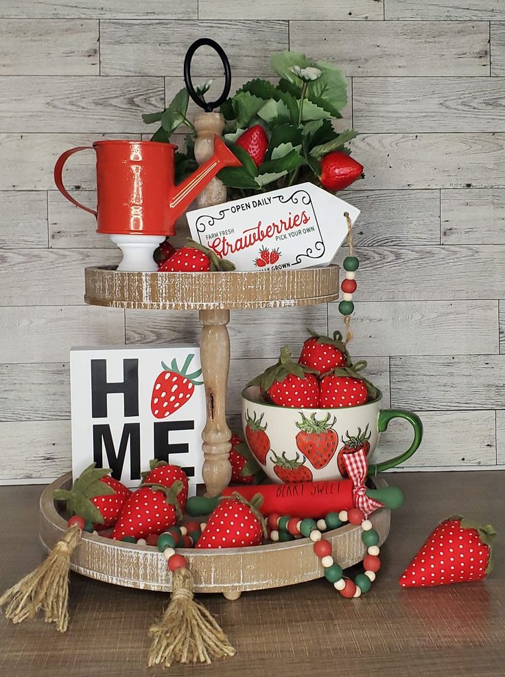 A two-tiered rustic wooden tray decorated with plush fabric strawberries, a "HOME" sign with a strawberry icon, a red watering can, and red and green beaded garlands.