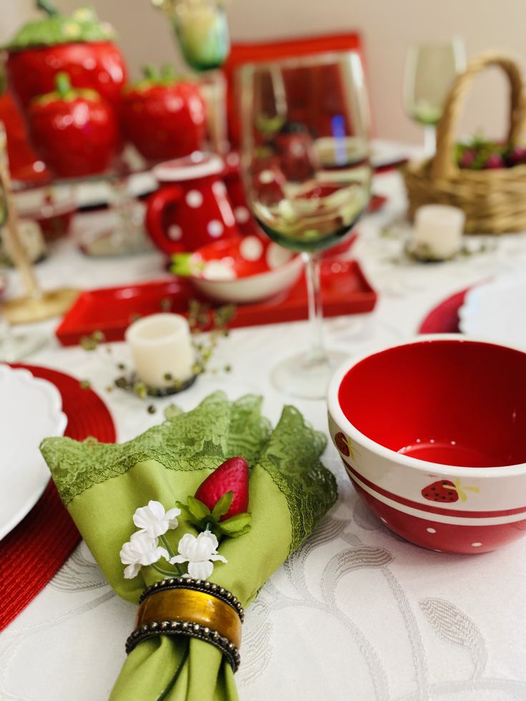 A close-up of a dining table set with a red and white polka dot bowl, a green lace napkin with a faux strawberry accent, and white dinnerware on a white tablecloth.