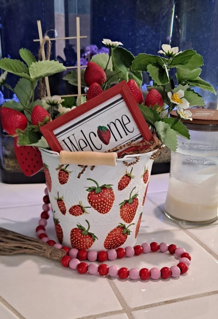 A white bucket with strawberry decor patterns filled with faux berries and a welcome sign.