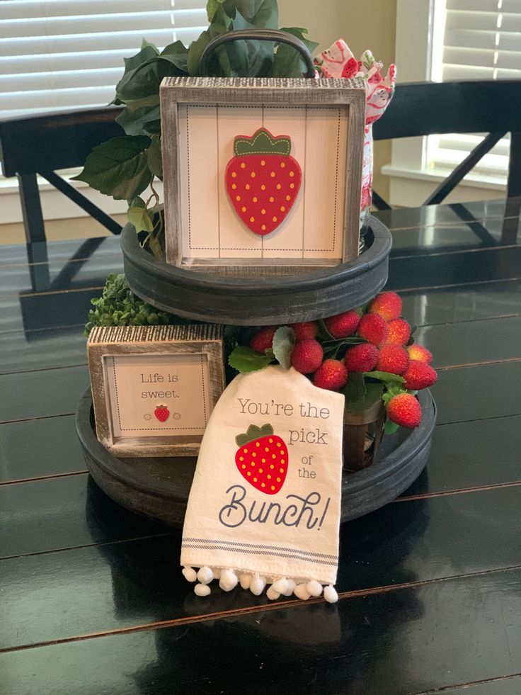 A two-tiered black tray on a dark wood table featuring strawberry-themed wooden signs, a white decorative towel with a strawberry graphic, and faux red berries.