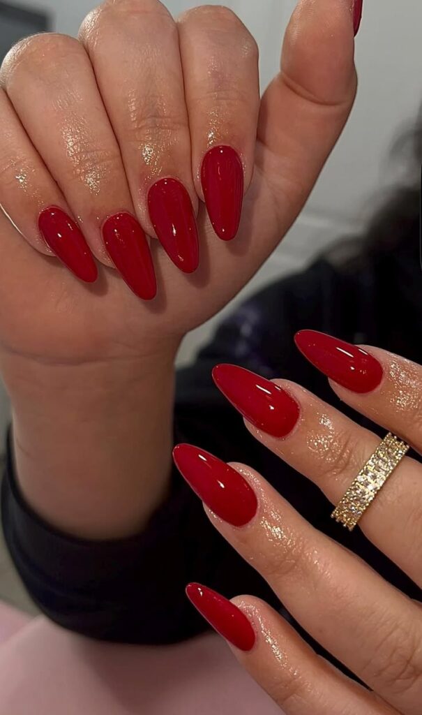 Two hands with long, almond-shaped deep red nails, one wearing a shimmering gold band.