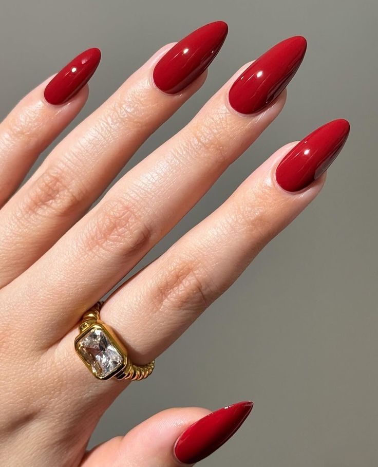 A hand with long, pointed ruby red nails wearing a large gold ring with a rectangular clear stone against a gray background.