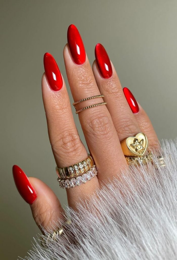 A hand with long glossy red almond nails wearing multiple gold rings and a white feathered sleeve.