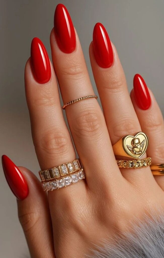 Close-up of long red oval nails with detailed gold jewelry, including a gold heart signet ring.