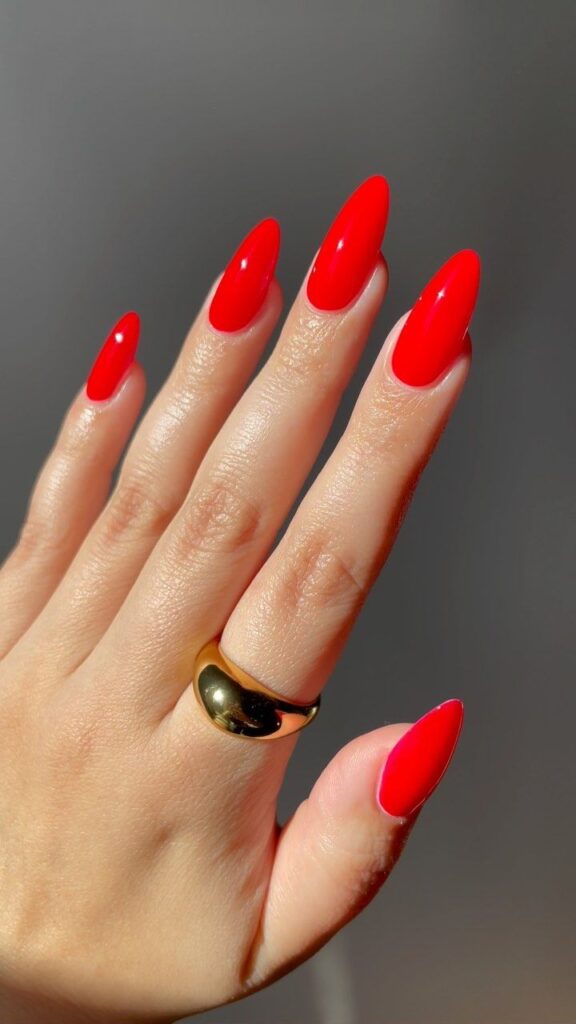 Tapered bright red nails on a hand wearing a single thick gold dome ring.