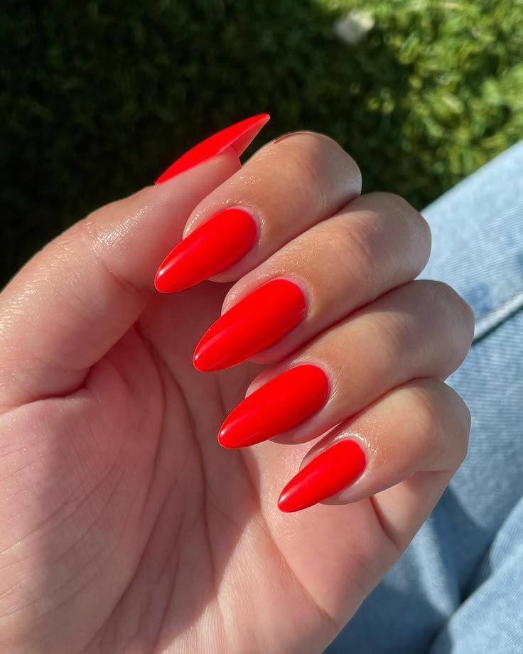 A hand with vibrant neon red almond nails held up against a sunny outdoor background of grass and denim jeans.