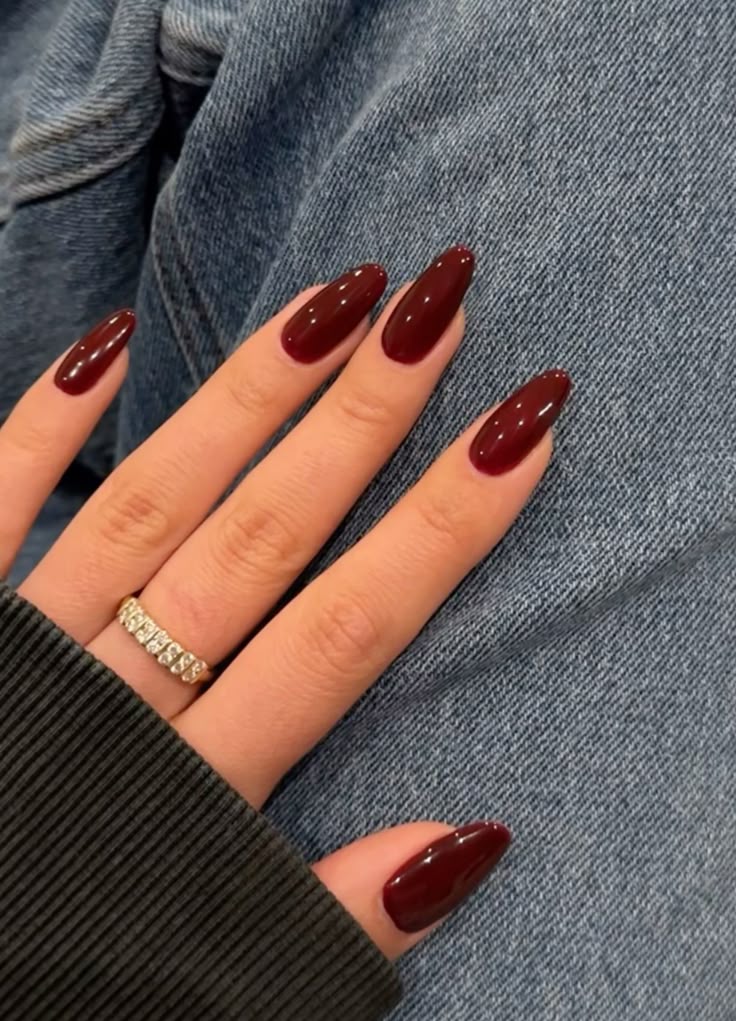Deep burgundy oval nails on a hand resting on denim jeans, wearing a gold and crystal eternity ring.