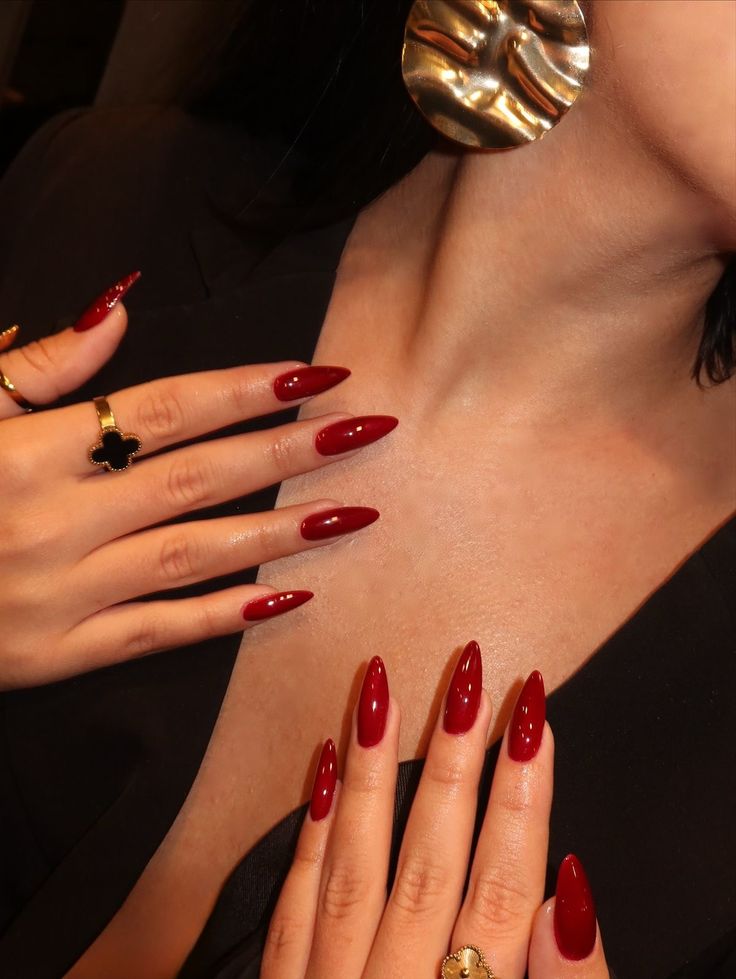 Two hands with long, glossy red pointed nails adorned with gold rings featuring black clover designs, and a large gold earring.