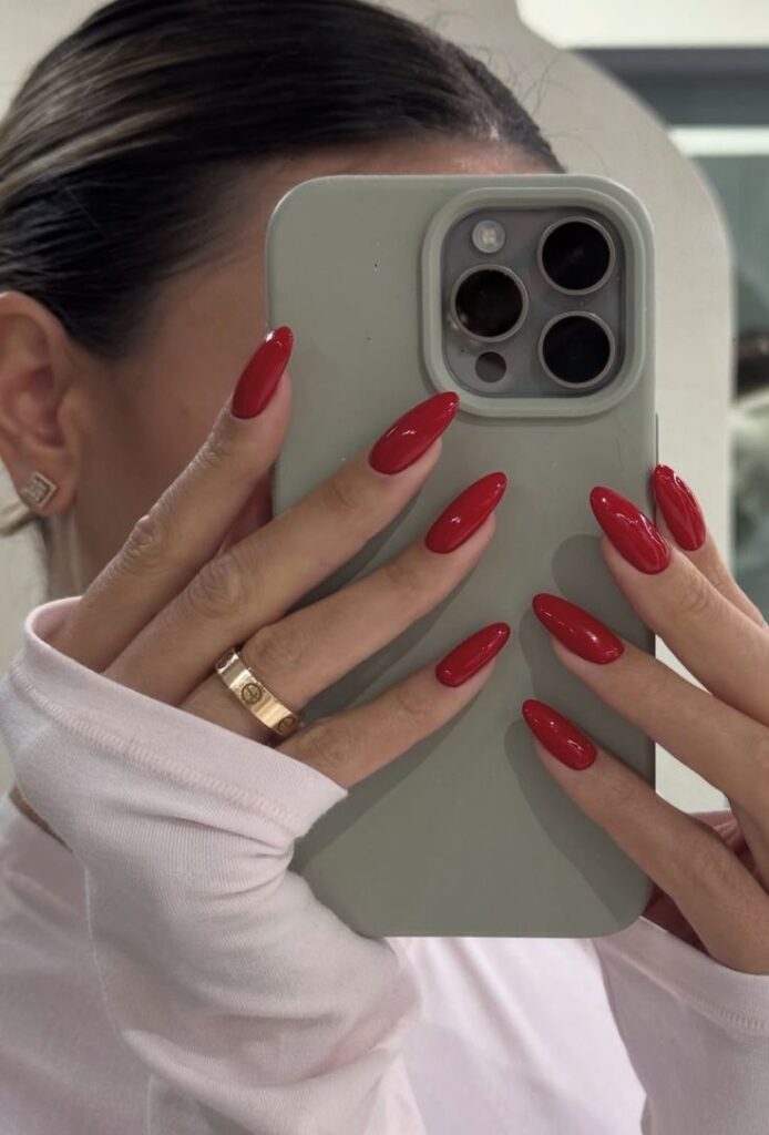A mirror selfie showing a person holding a green phone with long, glossy red almond-shaped nails and a gold band.