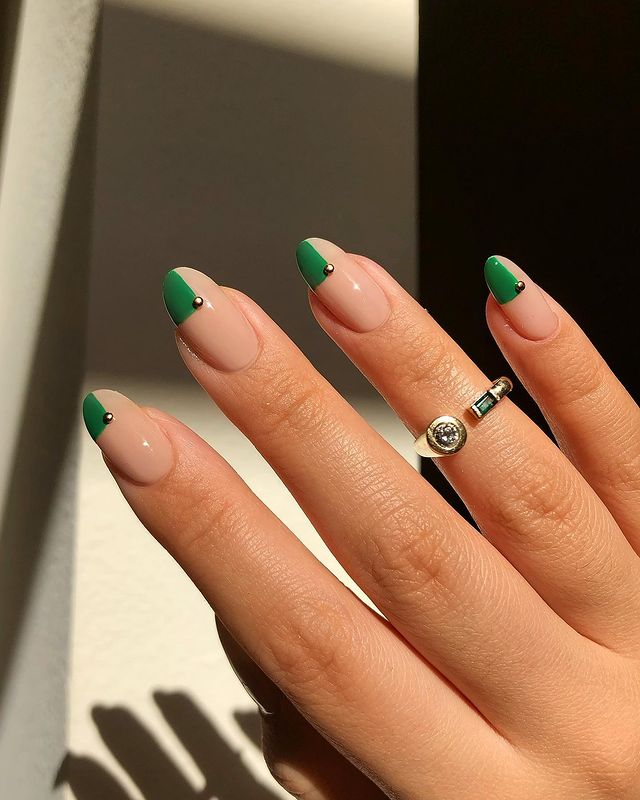 A hand with almond-shaped nails featuring a nude base and a green block accent on one side, decorated with a small gold dot.