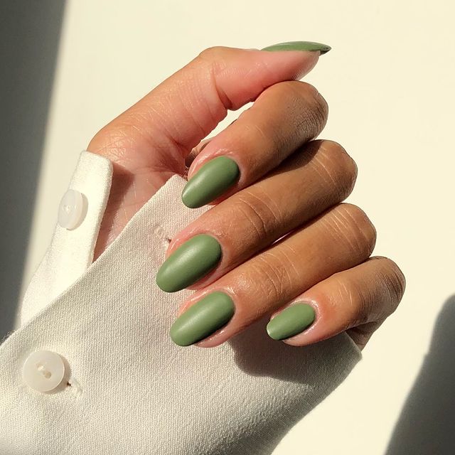 A close-up of a hand with almond-shaped nails painted in a solid, matte sage green color, wearing a white button-up shirt.