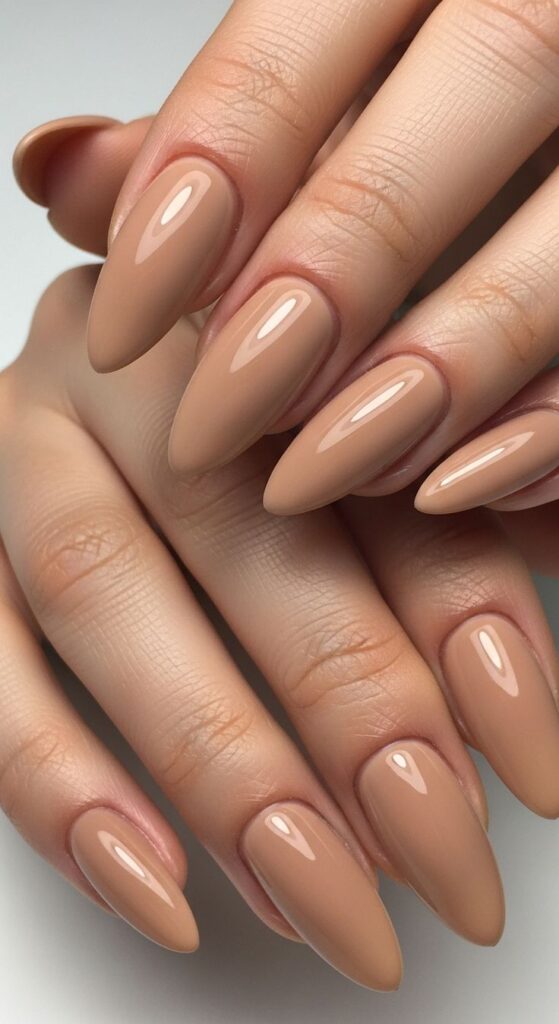 A close-up of two hands interlaced, featuring long, almond-shaped nails painted in a high-gloss, warm toffee caramel camel nude nail polish. The smooth, creamy texture of the polish complements the natural, warm undertones of the skin.