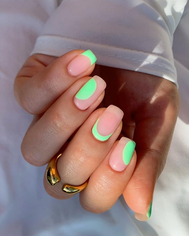 A close-up of a hand with short, square-shaped nails featuring a pink base and neon green half-moon accents, styled with a gold ring.